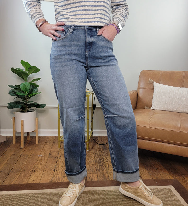 Indoors on a wooden floor, someone wears Risen’s Mira Cropped Barrel Jeans, a striped long-sleeve shirt, and beige sneakers. A plant in a white pot and a tan couch with a white pillow are in the background.
