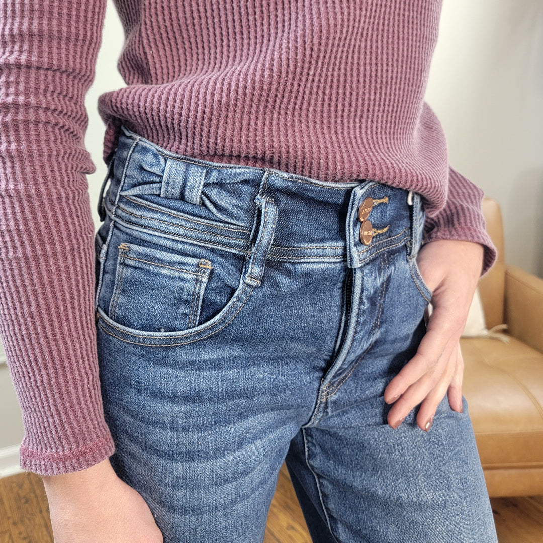 Someone stands indoors with one hand in their pocket, wearing a textured mauve long-sleeve top and Geneva Wide Leg Jeans from Risen with Tummy Control. A tan armchair is visible in the background.
