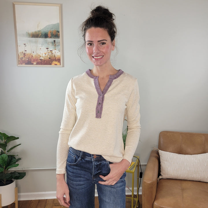 A woman with brown hair in a bun, wearing the Staccato Naomi Long Sleeve Henley and blue jeans, smiles in a cozy living room featuring a brown leather chair, houseplant, and a nature painting on the wall.