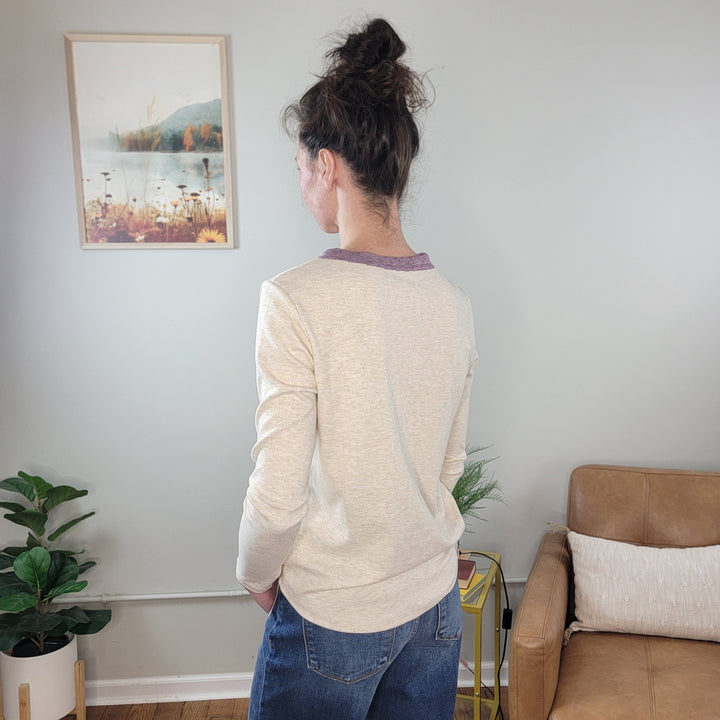 A person with brown hair in a messy bun faces away in a cozy living room, wearing the Staccato Naomi Long Sleeve Henley and blue jeans—an ideal outfit for cold weather amid framed art, plants, and a tan sofa.
