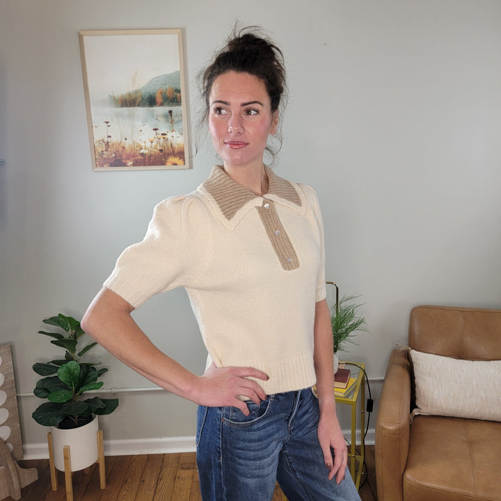 A woman with dark hair in a bun wears the Allie Rose Luna Short Sleeve Sweater and blue jeans. Indoors, she stands with one hand on her hip, surrounded by plants, a painting, and a tan chair.