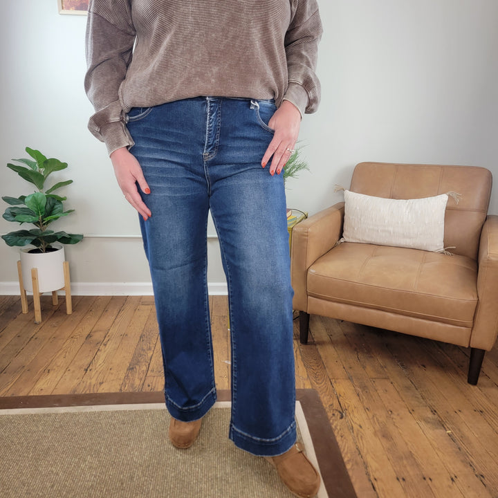A person wearing Sophie Straight Jeans from Risen with Tummy Control by Cosmos + Clover Boutique, a taupe sweater, and brown shoes stands on a wooden floor next to a tan armchair and potted plant in a cozy, well-lit room.