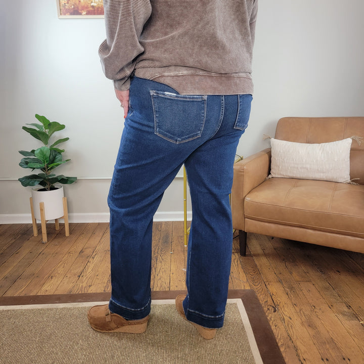 A person wearing Sophie Straight Jeans from Risen with Tummy Control by Cosmos + Clover Boutique, a brown striped top, and brown shoes stands on a wooden floor near a tan armchair and green potted plant in a cozy room.
