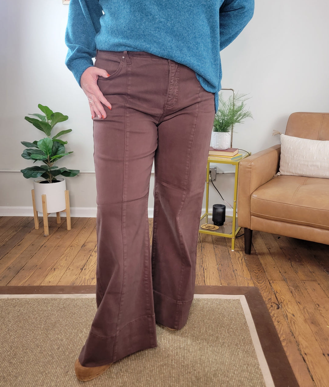 A person stands indoors, their face not visible, wearing Risen’s Kayla Chocolate Brown Wide Leg Jeans with Tummy Control. They are on a wood floor next to a tan chair, green plants, and a small side table.
