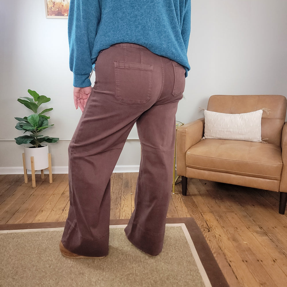 A person wearing Risen Kayla Chocolate Brown Wide Leg Jeans with Tummy Control and a blue sweater stands on a wooden floor, back to the camera. A tan armchair with a white pillow and a green potted plant are in the background.