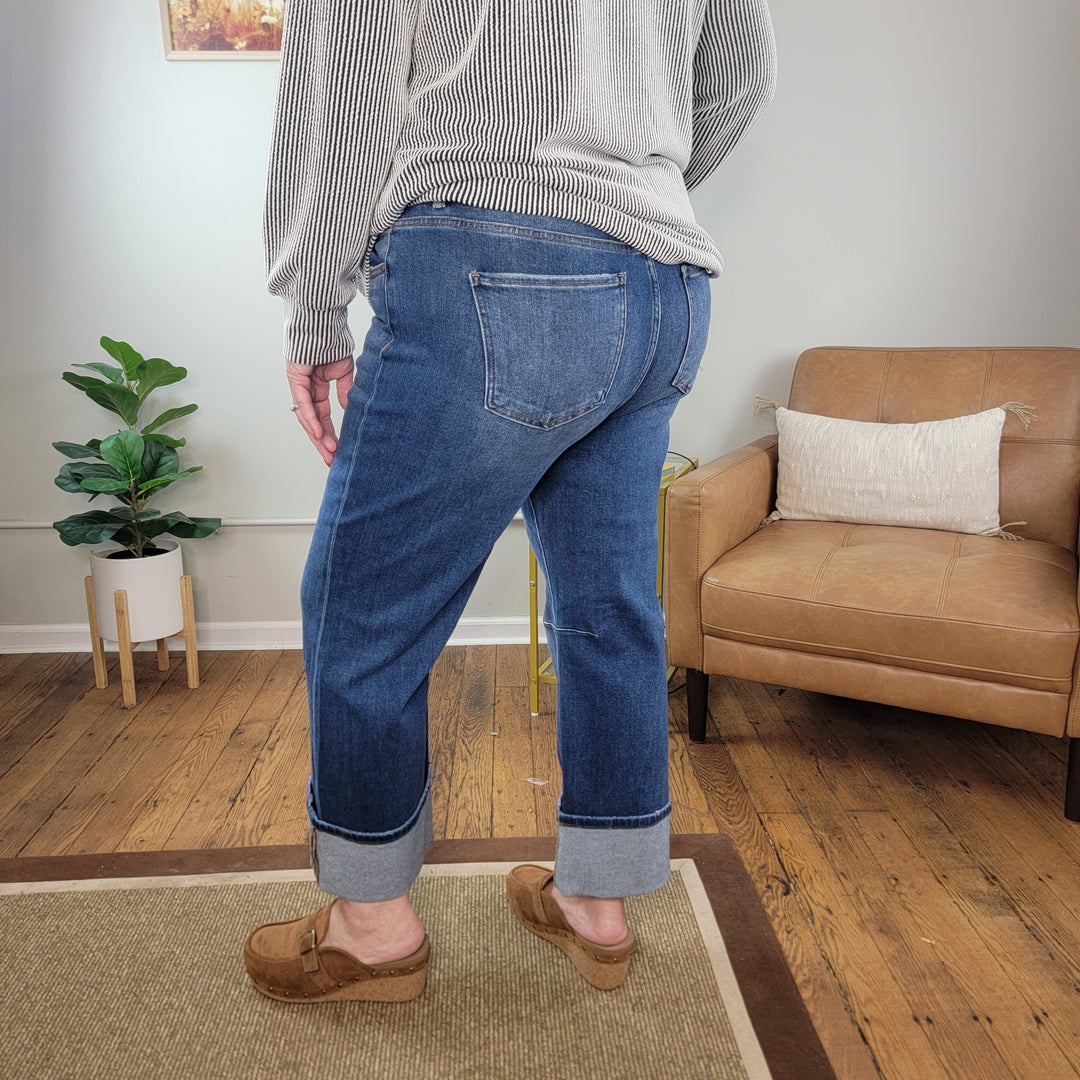 A person stands on a hardwood floor near a tan armchair and green plant, wearing Risen’s Lakyn Straight Jeans, a gray sweater, and brown shoes in a modern living room.