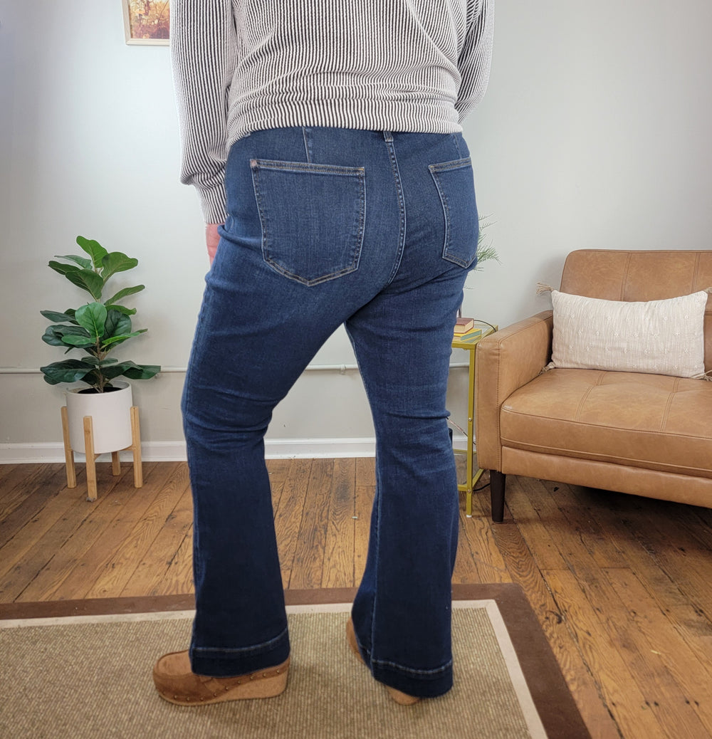 Someone wearing Reya Flares from Judy Blue with a striped top and brown shoes stands indoors on a wooden floor near a tan chair, green plant, and small table. The photo is taken from behind to highlight the jeans' fit.
