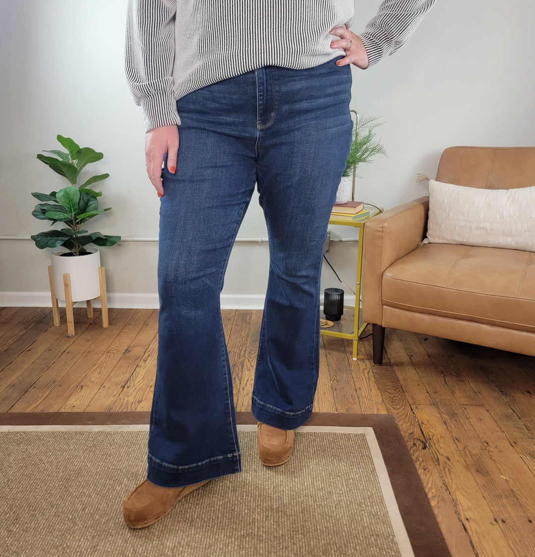 A person stands indoors on a wooden floor wearing Judy Blue’s Reya Flares, a striped sweater, and brown shoes. In the background, there’s part of a beige chair, a plant, and a yellow side table.