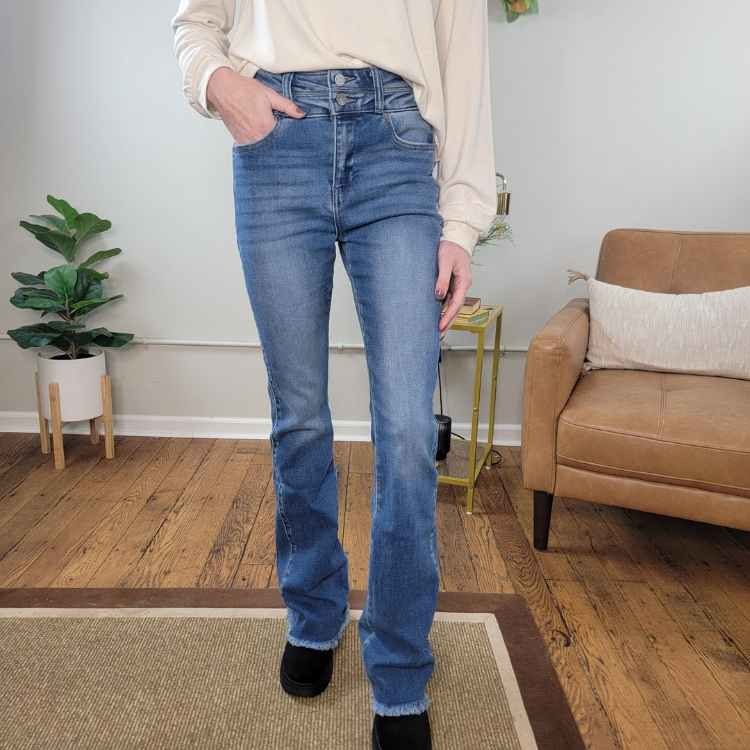 A person wearing KanCan Vanessa Side Panel Bootcut Jeans and a light beige long-sleeve top stands indoors on a wood floor, with a tan armchair, a pillow, and a green potted plant in the background.
