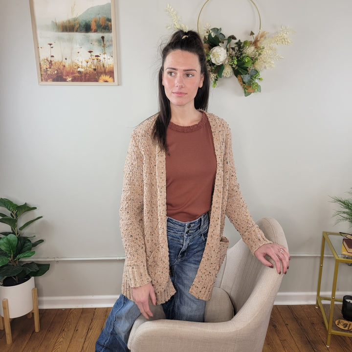 A woman with long dark hair, in a rust shirt, blue jeans, and the Staccato Parker Popcorn Open Front Cardigan, stands by a light chair in a cozy room with wood floors, plants, a floral wreath, and framed art.