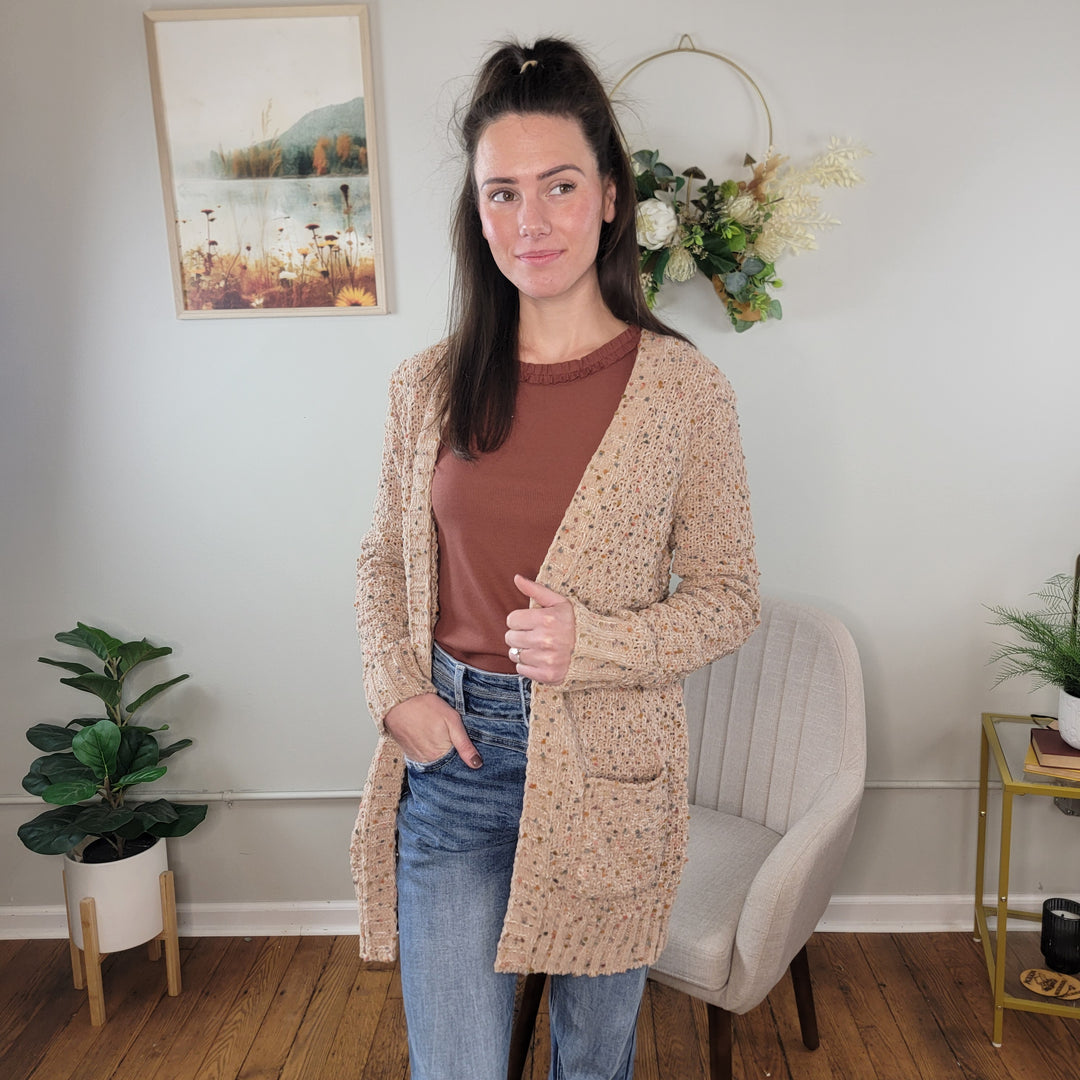 A woman with long brown hair wears the Staccato Parker Popcorn Open Front Cardigan in beige over a brown top and blue jeans. She stands in a warm room decorated with plants, a chair, framed nature art, and a floral wreath on the wall.