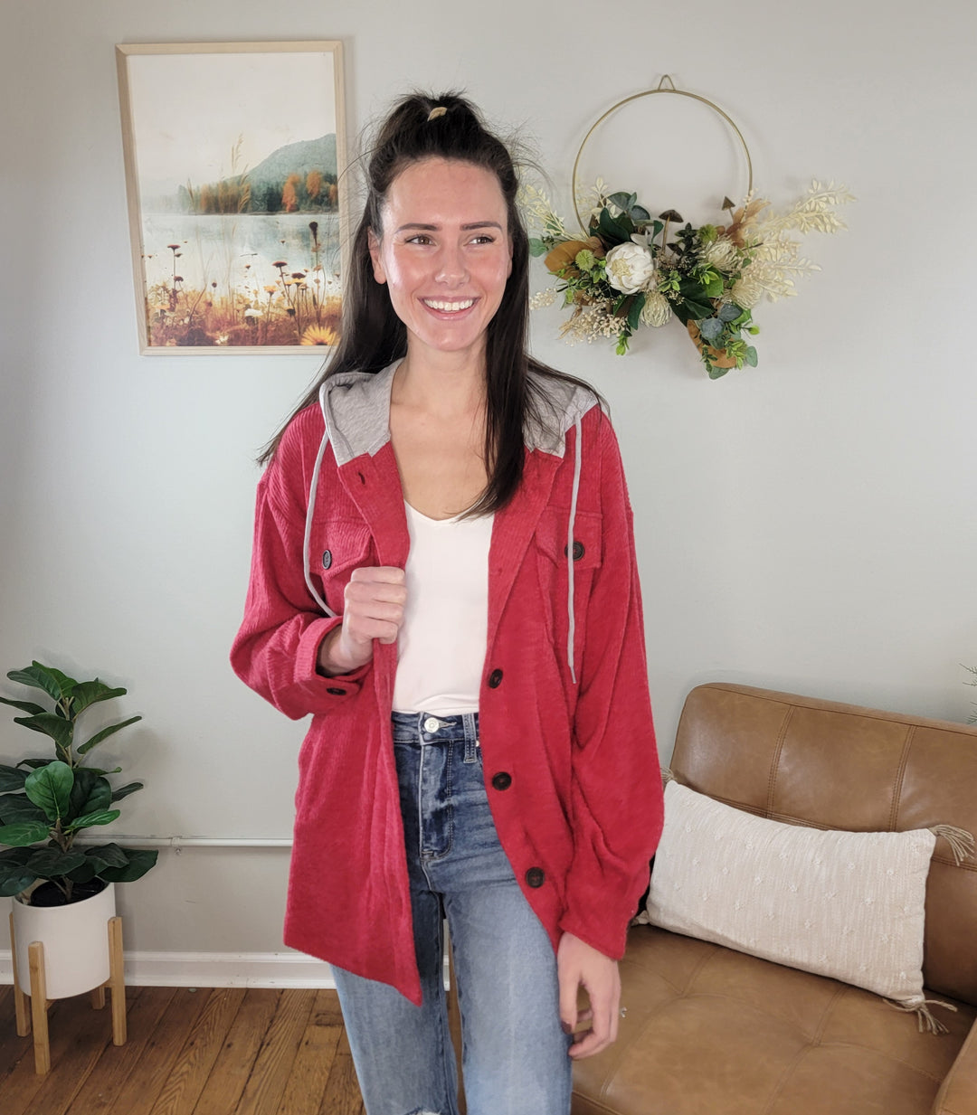 A woman with long brown hair smiles in a cozy living room, wearing the ZENANA Kysa Button Front Shacket with Hood over a white top and blue jeans, surrounded by plants, a wall wreath, and framed art.