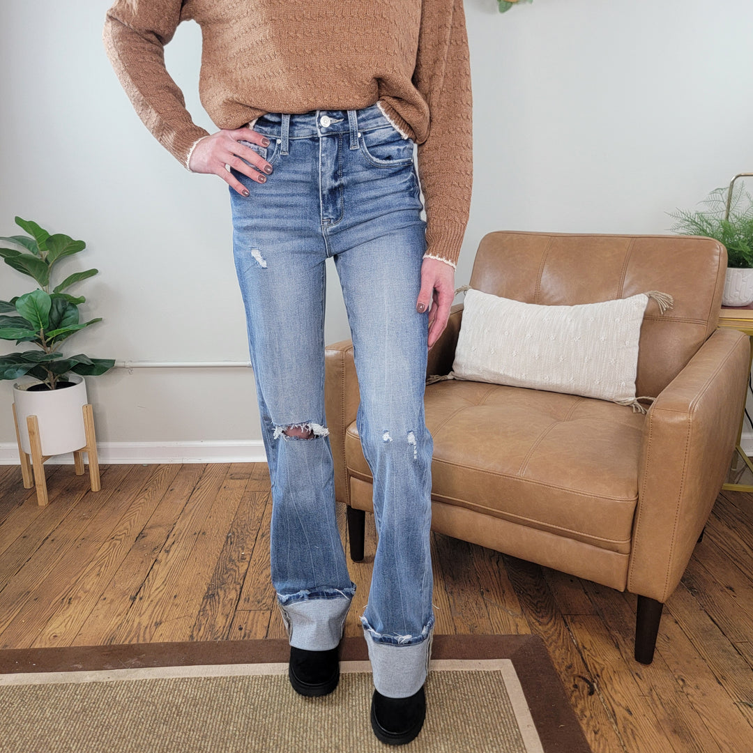 A person stands indoors on a wooden floor, wearing a textured brown sweater, black shoes, and the Jennifer Wide Leg Flare jeans from Petra153 with cuffed hems and distressing. Behind them is a tan armchair with a white pillow and green potted plants.