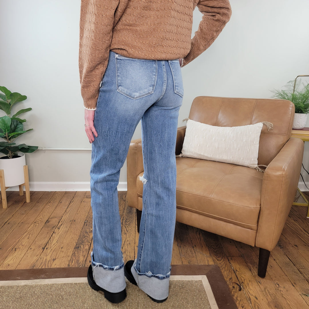 Wearing a brown sweater, black shoes, and Petra153’s Jennifer Wide Leg Flare jeans, a person stands on hardwood near a tan armchair with white pillow and small plant against a white wall. Photo is taken from behind.