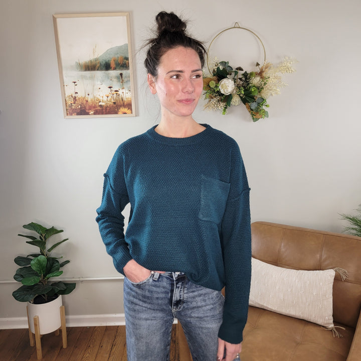 A woman with dark hair in a bun, smiling and looking to the side, wears the Staccato Taelor Knit Sweater with Chest Pocket and jeans. She stands in a cozy living room with plants, wall art, a leather couch, and a pillow.
