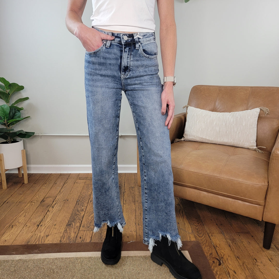 Someone wearing Petra153’s Diana Slim Wide Leg Jeans, a white t-shirt, and black boots stands on a wooden floor indoors beside a tan chair and potted plant.