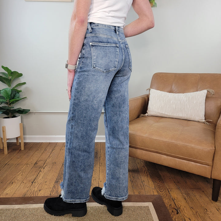 A person in a white shirt and Petra153's Diana Slim Wide Leg Jeans stands on a rug in a living room with wooden floors, a tan armchair with a pillow, and a green potted plant in the background.