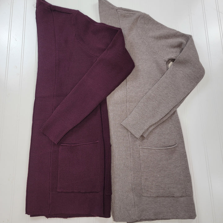 Two Staccato Ashley Cardigans are displayed flat on a white surface—one in deep plum, the other in light beige. Both feature long sleeves, ribbed knit fabric, open front design, front pockets, and a cozy style.