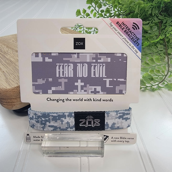 A ZOX Bracelets band from ZOX, featuring light blue and gray patterns, is shown in packaging labeled FEAR NO EVIL. Made from recycled water bottles, this interactive bracelet reveals a new Bible verse with each tap; a leafy plant sits behind.