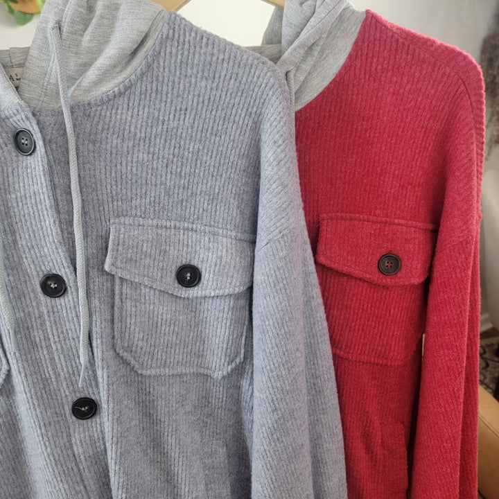 Two ZENANA Kysa Button Front Shackets with Hoods hang side by side on wooden hangers—one in heather grey and one in red—each featuring black buttons and front pockets, perfect as cozy layering pieces.
