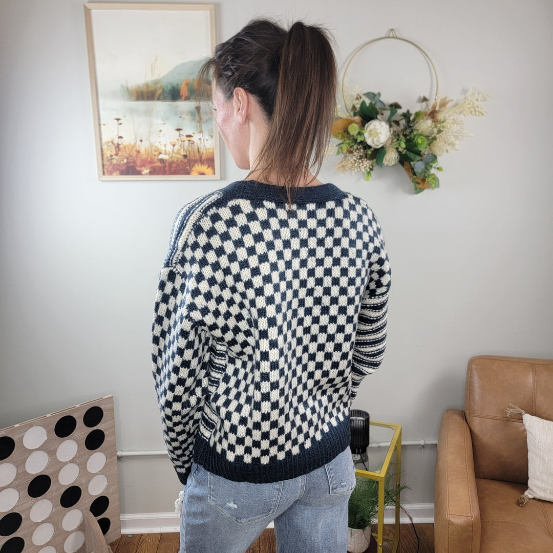 A person with a ponytail, wearing the HYFVE, Inc Gemma Button Down Sweater and light jeans, stands indoors facing away. The room includes wall art, a floral wreath, a tan chair, and a board game on the floor.