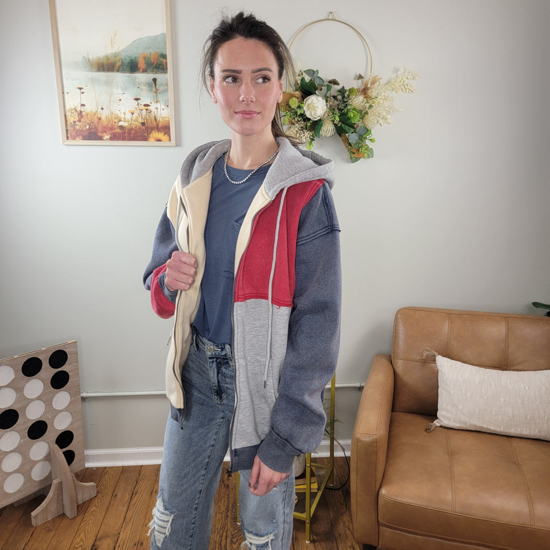A woman wearing the HYFVE, Inc Blythe Color Block Zip-Up and ripped jeans stands in a cozy living room with a tan armchair, wall-mounted floral arrangement, and framed art in the background.