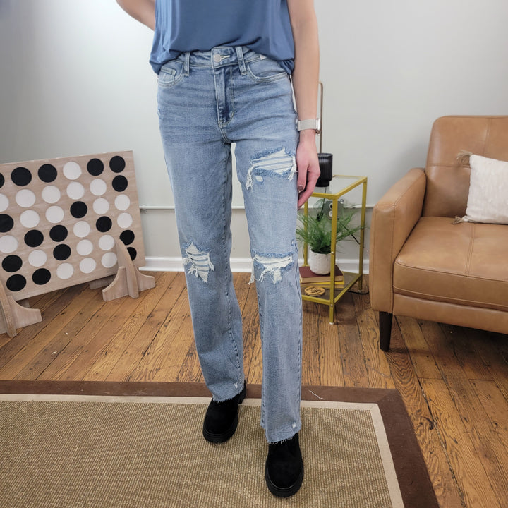Someone indoors models the Judy Blue Lilly 90's Straight Jeans—a light wash, distressed denim—paired with black shoes. Only their lower torso and legs show, set against a tan armchair, plant on a shelf, and polka dot board.