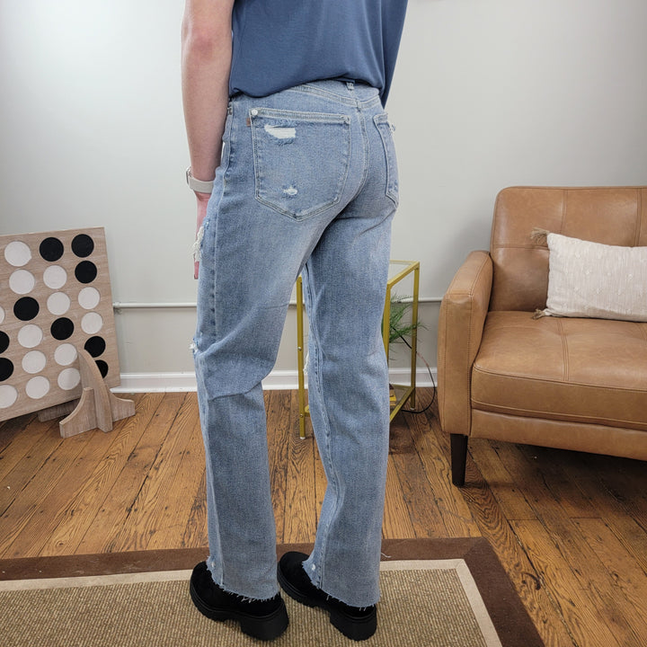 Indoors, a person faces away on a rug, wearing Judy Blue’s Lilly 90’s Straight Jeans in light wash, black shoes, and a blue shirt. A brown leather chair and large Connect Four game are visible in the background.