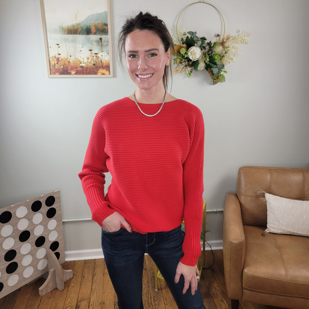 A woman smiles at the camera indoors wearing the Allie Rose Amelia Ribbed Sweater in red with jeans; behind her are a brown armchair with a white pillow, wall art, a floral wreath, and a large Connect Four game on the floor.
