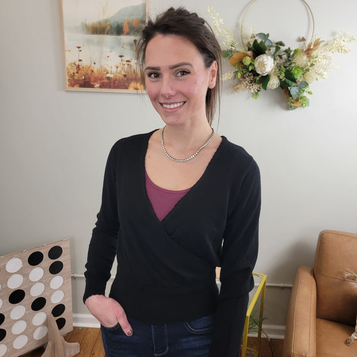 A woman with dark hair in a ponytail, wearing Allie Rose's Loxly cross-over sweater and a pearl necklace, smiles indoors. Behind her are a tan armchair, framed art, and a large wall wreath.
