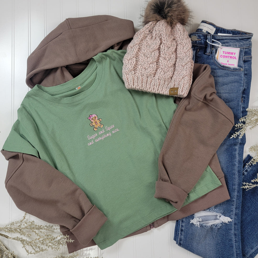 Flat lay of a casual outfit featuring the Mono B Sugar and Spice Cropped Tee, layered under a brown sweatshirt, with a light pink knit beanie with faux fur pompom, and blue skinny jeans labeled for tummy control.