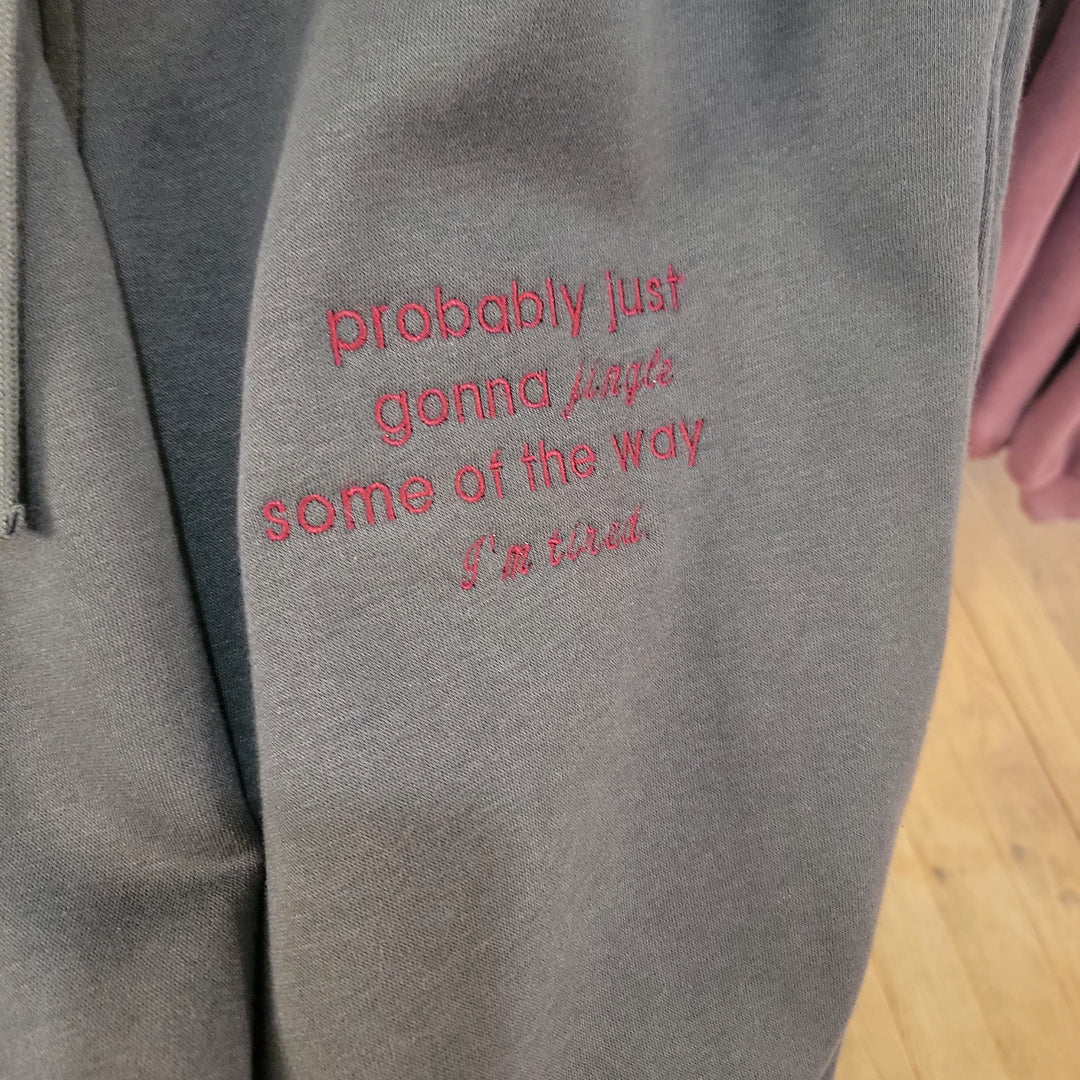 A close-up of Rae Mode's "I'm Tired" Embroidered Joggers in gray, featuring red text that reads "probably just gonna jingle some of the way I’m tired," shown against a wooden floor with another garment in the background.