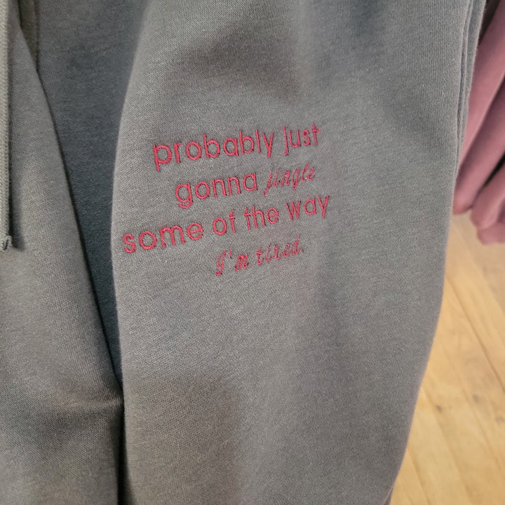 A close-up of Rae Mode's "I'm Tired" Embroidered Joggers in gray, featuring red text that reads "probably just gonna jingle some of the way I’m tired," shown against a wooden floor with another garment in the background.