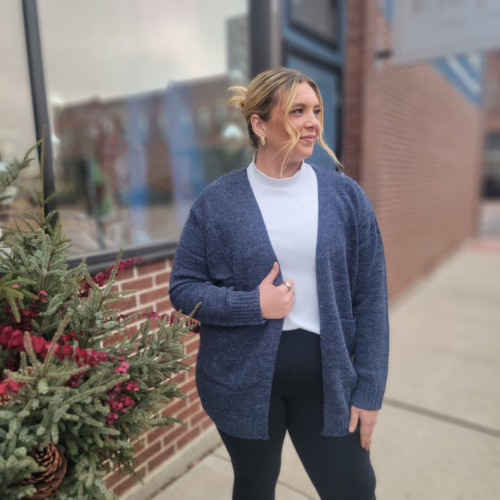 A woman stands outside by a building, wearing the ZENANA Daciana Open Front Knit Cardigan in navy over a white top and black pants, smiling gently with a festive red-berried plant in the foreground.