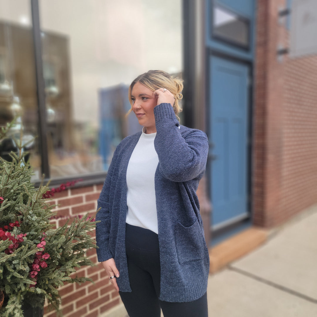 A woman with blond hair stands by a brick wall and window, wearing the ZENANA Daciana Open Front Knit Cardigan in navy over a white shirt and black pants. She touches her hair and looks to the side, with plants beside her.