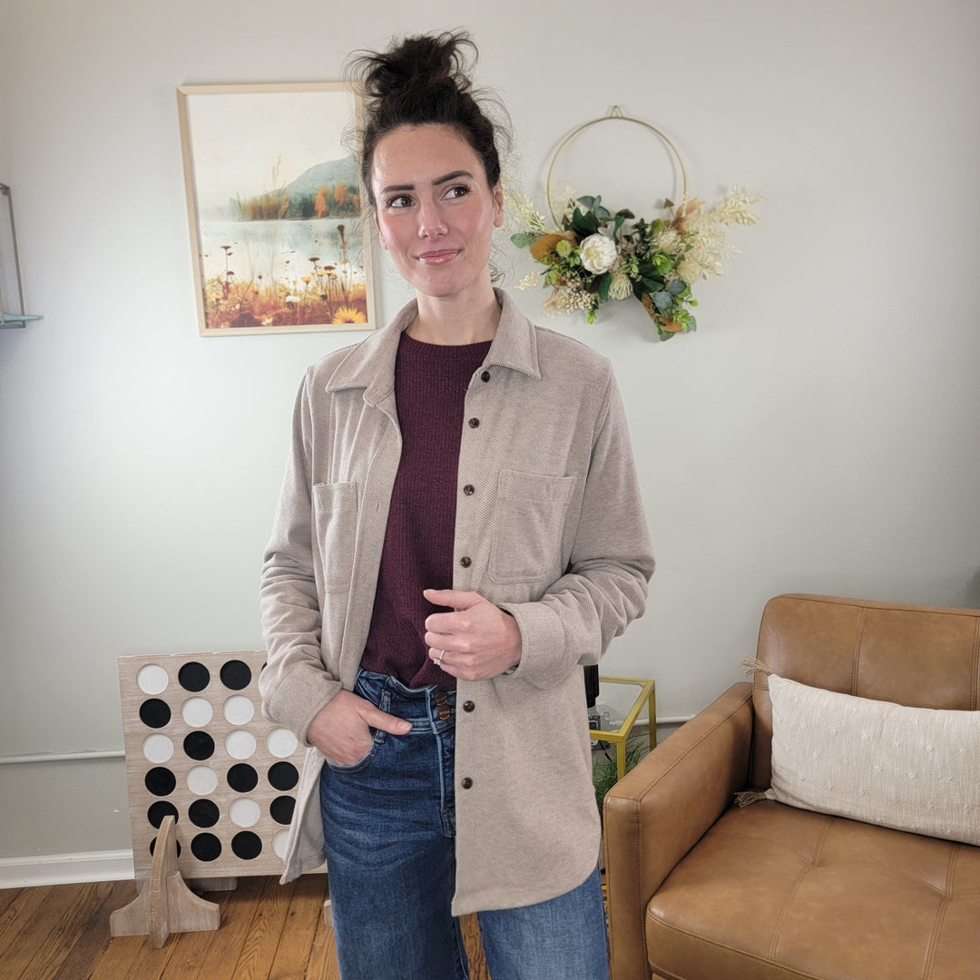 A woman with dark hair in a messy bun stands indoors, smiling in the Panache Callie Tan Flannel Button Down Shacket over a maroon top and blue jeans, one hand in her pocket. A brown chair and wall decor are visible behind her.