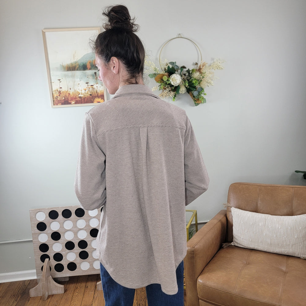 A person with long dark hair in a bun, wearing the Panache Callie Tan Flannel Button Down Shacket and jeans, stands with their back to the camera in a living room featuring wall art, a floral wreath, a leather chair, and a large Connect Four game.