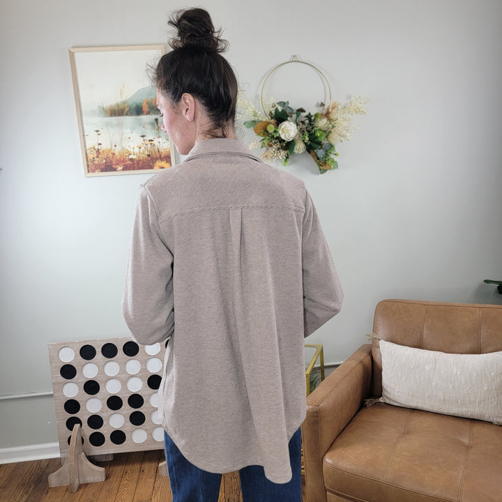 A person with long dark hair in a bun, wearing the Panache Callie Tan Flannel Button Down Shacket and jeans, stands with their back to the camera in a living room featuring wall art, a floral wreath, a leather chair, and a large Connect Four game.