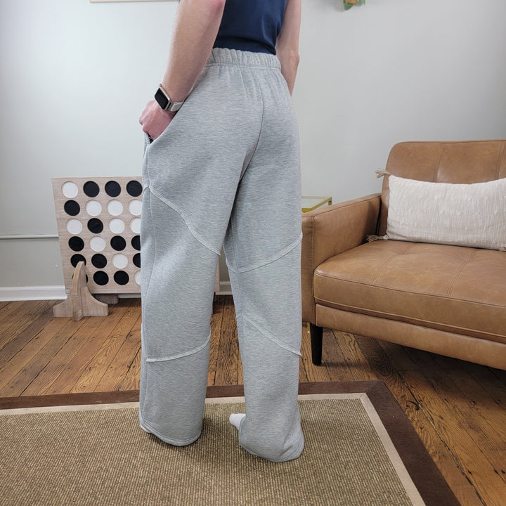 A person wearing HYFVE, Inc's Sloan Wide Leg Pants in light gray stands on a rug in a living room with wooden floors, a tan couch, white pillow, and a large wall game board visible in the background.