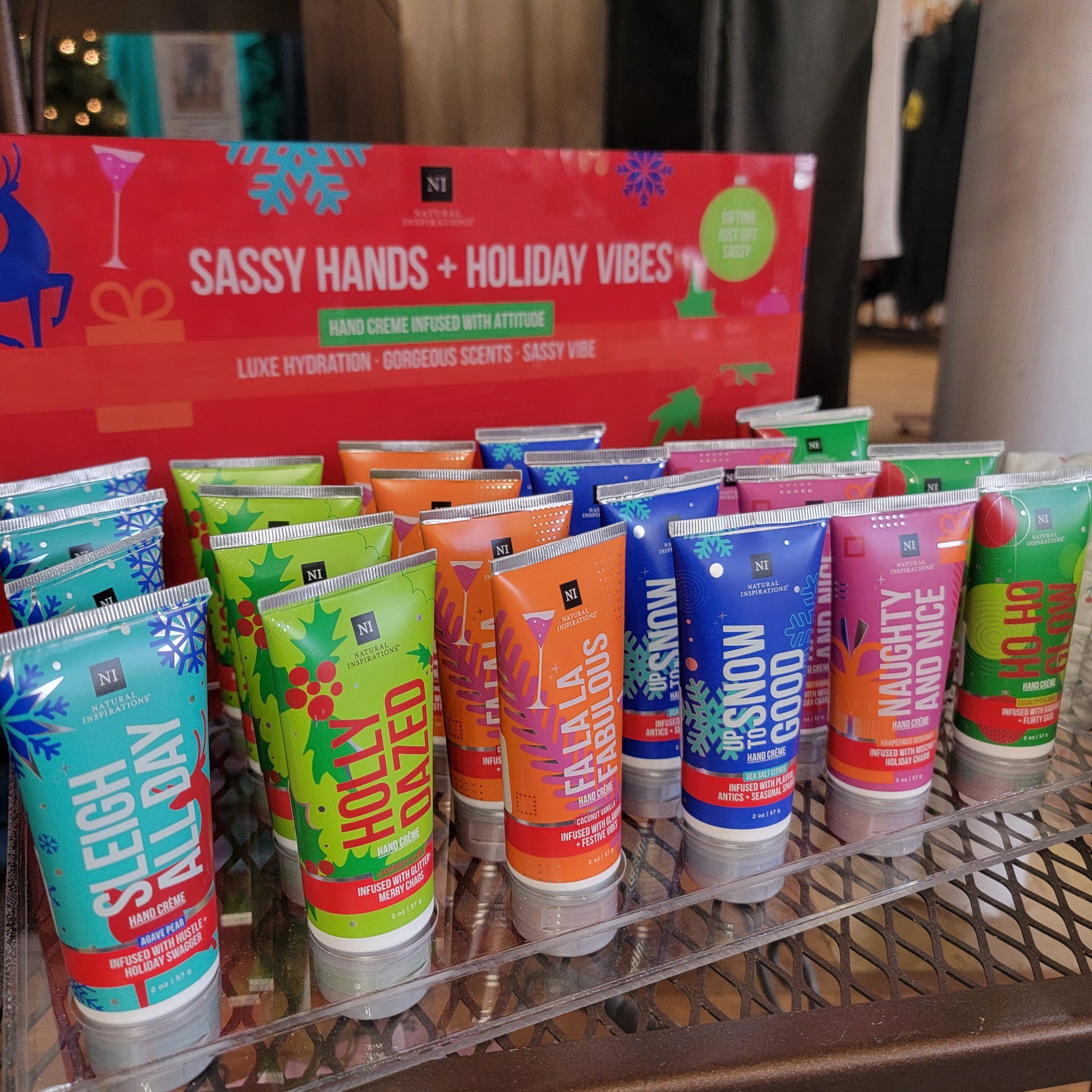 A display of colorful 2 oz Natural Inspirations Hand Lotions by Natural Inspirations, arranged in rows before a red "Sassy Hands + Holiday Vibes" sign, highlights a festive holiday assortment with cheerful seasonal graphics.