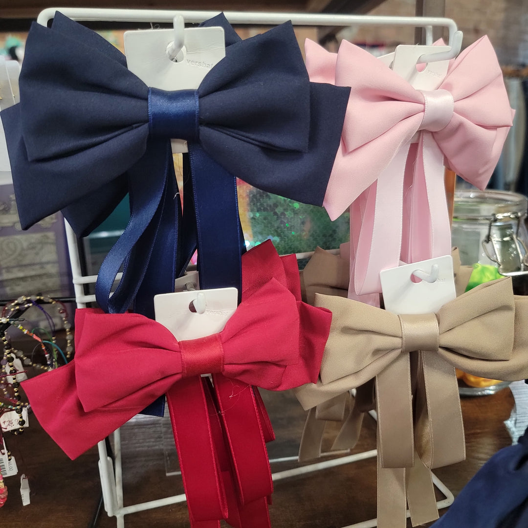 Hair Bows— large assorted bows in navy blue, light pink, red, and beige—are displayed on white cards hanging from a metal rack, with colorful accessories and bracelets in the background.