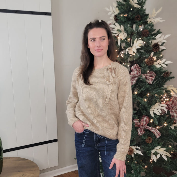 A woman with long brown hair wears the Hem &amp; Thread Alayla Tie Neck Knit Sweater and blue jeans as she stands indoors beside a decorated Christmas tree with ornaments, pinecones, and plaid ribbons, smiling softly to her left.