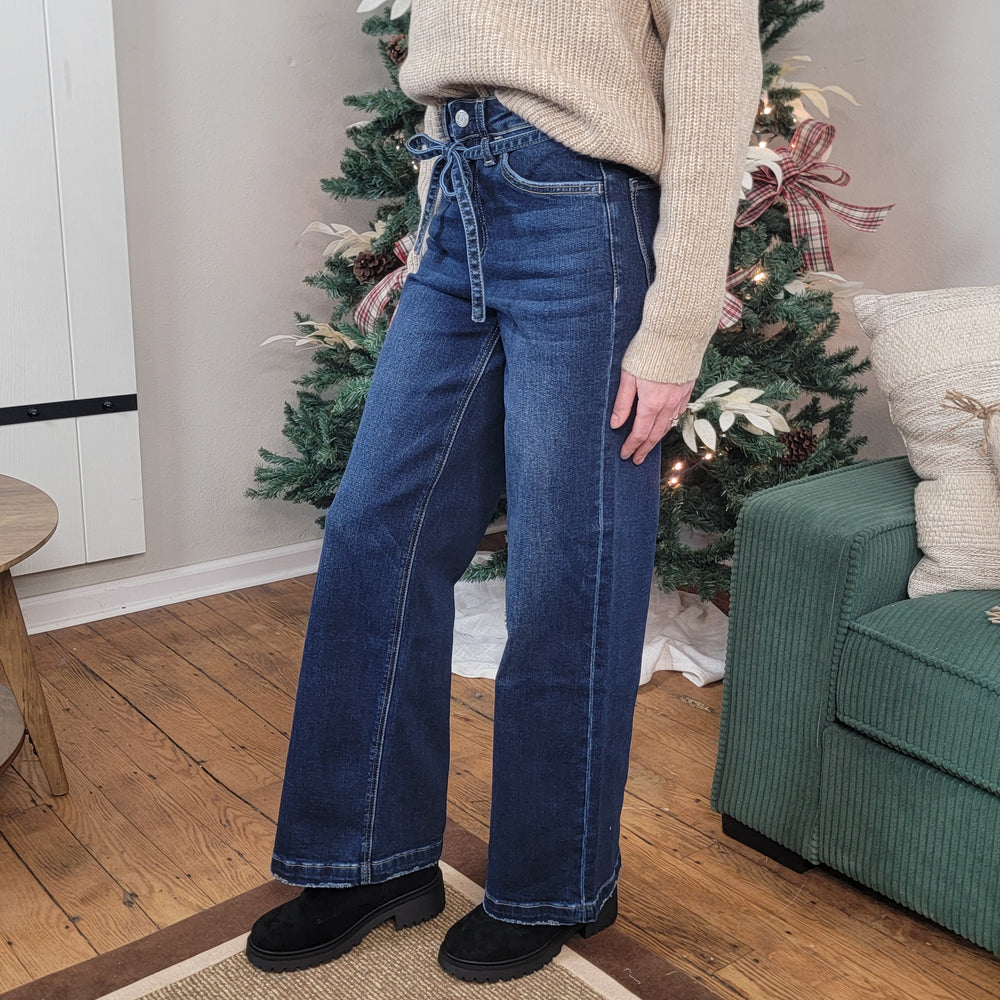 Indoors on a wooden floor, a person wears Vervet's Astonishment Wide Leg Jeans with a beige sweater and black shoes. A decorated Christmas tree and part of a green sofa appear in the background.