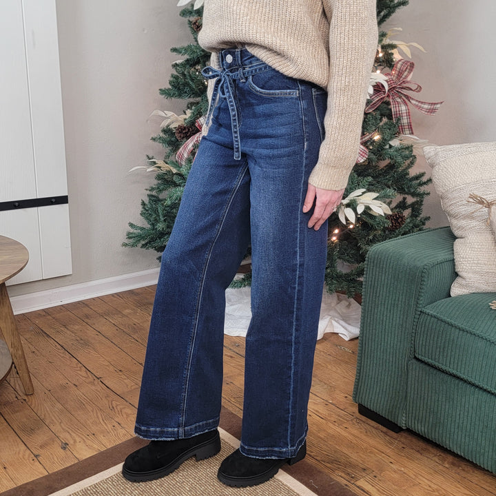A person stands indoors on a wooden floor, wearing Astonishment High Rise Wide Leg Jeans from Vervet by Flying Monkey, black shoes, and a beige sweater. A decorated Christmas tree and a green couch are in the background.