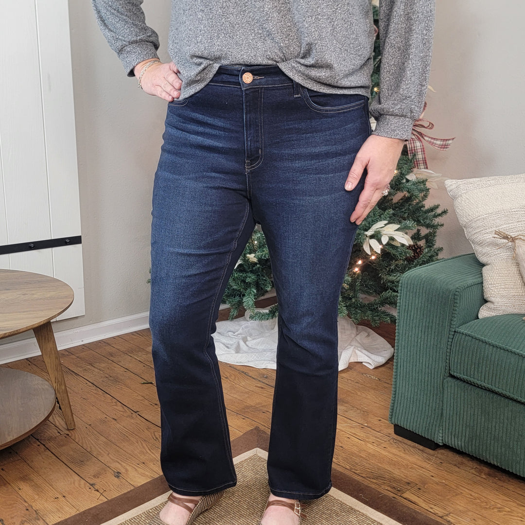 A person wearing Vervet's Scottie Bootcut Jeans in dark wash comfort stretch denim and a gray long-sleeve top stands indoors on a wooden floor near a green couch and Christmas tree. Their face is not shown.