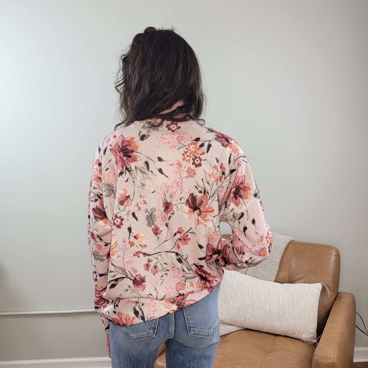 A person with dark hair, wearing the Fate Selma Floral Sweater—a pink crewneck with watercolor florals—and blue jeans stands with their back to the camera in a room with beige walls and a tan chair topped by a white pillow.