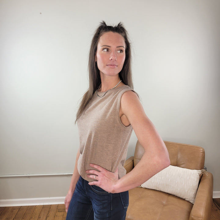 A woman with long brown hair stands indoors, wearing the Staccato Helena Sleeveless Sweater and dark jeans. She poses with one hand on her hip beside a tan leather chair with a white pillow against a light-colored wall.