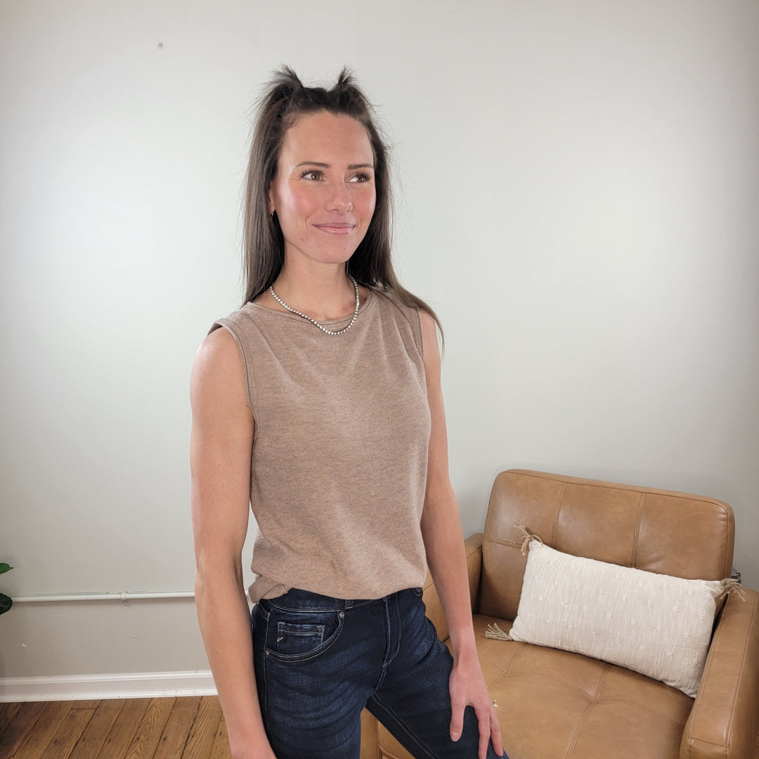 A woman with long brown hair, wearing the Staccato Helena Sleeveless Sweater and dark jeans, stands indoors by a tan leather chair with a white pillow. The room has light walls and wooden floors.