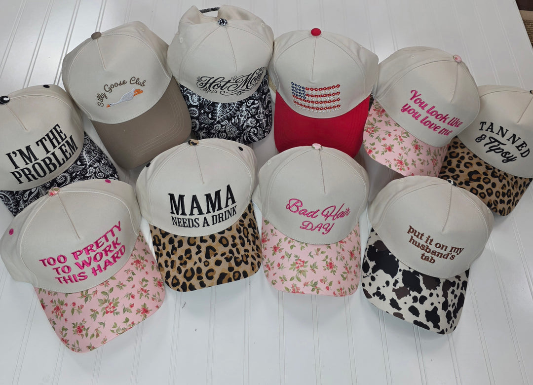 A set of twelve ZENANA Embroidered Hats is displayed on a white surface, each featuring a unique design with humorous embroidered phrases. The collection showcases various patterns—including leopard, floral, bandana, and cow print—making these miscellaneous caps perfect for anyone who enjoys hats with witty sayings.