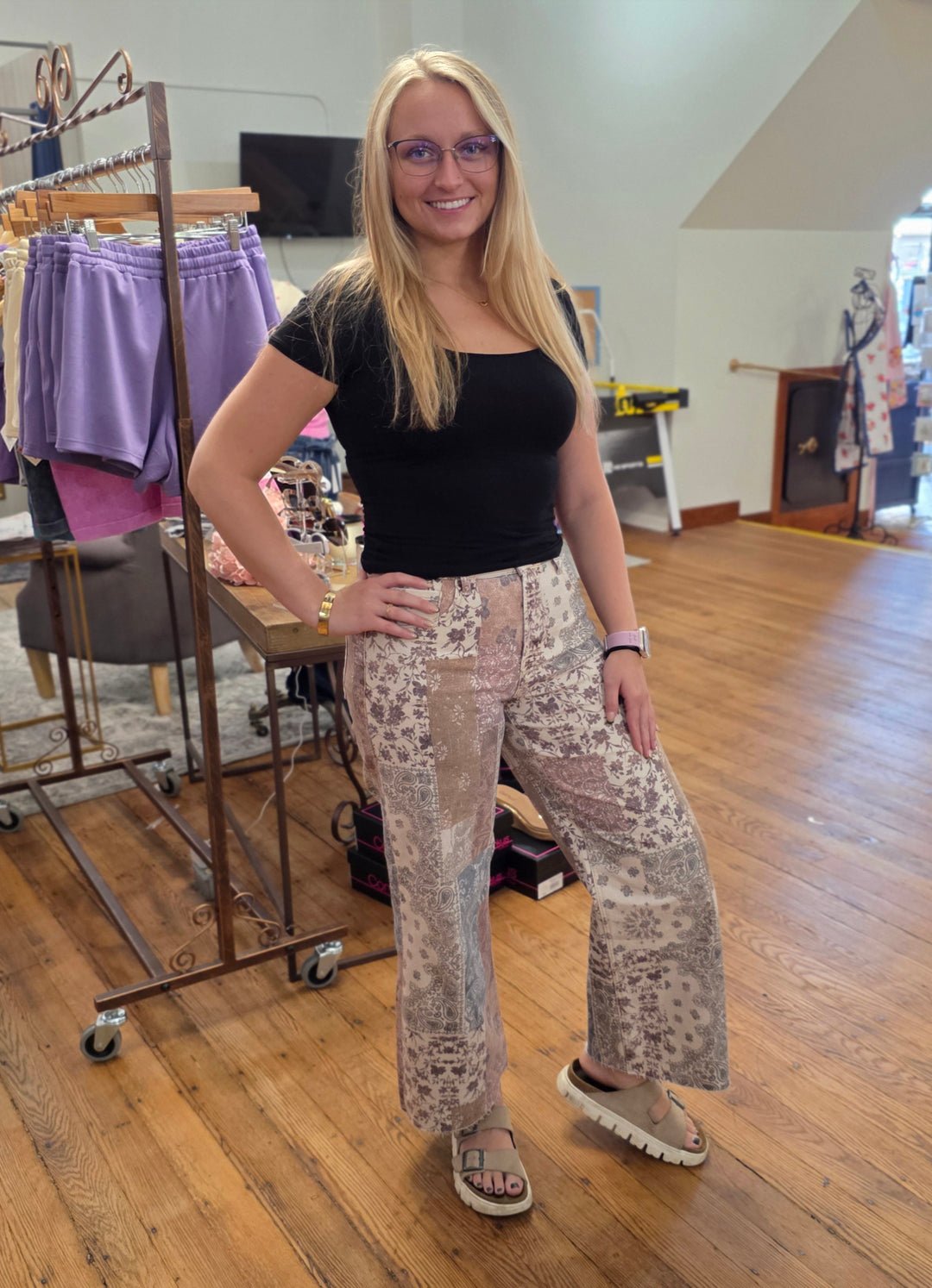 A woman in a black top and patterned pants stands smiling in a boutique, surrounded by clothing racks and wooden floors. The inviting atmosphere highlights an array of fashion-forward pieces like the Aven High Rise Ankle Wide Leg Jeans from Risen—crafted with stretch denim—for stylish, comfortable updates to any wardrobe.